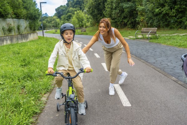 Happy mother running alongside her son, guiding him as he learns to ride a bicycle with training wheels in a vibrant park, soaking up the joy of a sunny day outdoors