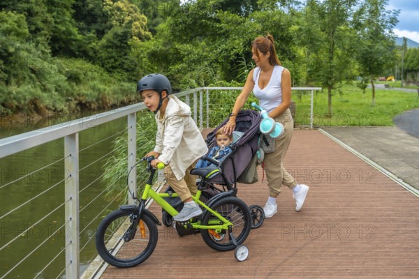 Young mother pushing a stroller with a baby inside while watching her other son riding a bicycle near a river in a park, enjoying a sunny day together