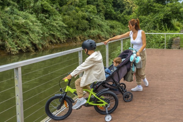 Mother walking with her baby in a stroller and her older son riding a bicycle on a wooden walkway next to a river, enjoying a sunny summer day surrounded by lush nature
