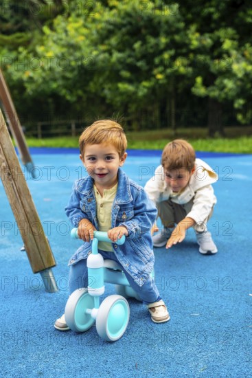 Happy toddler boy riding a balance bike on a playground with his older brother cheering him on, promoting childhood activities, outdoor play, and sibling bonding