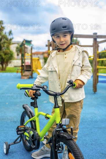 Young boy wearing a helmet standing and holding the handlebars of his bicycle with training wheels in a playground with a blue rubberized floor