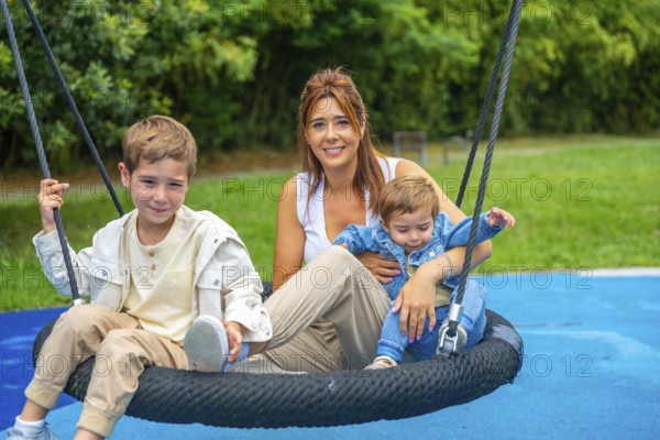 Happy family enjoying a sunny day at the playground, the mother is swinging with her two children on a nest swing, having fun together outdoors