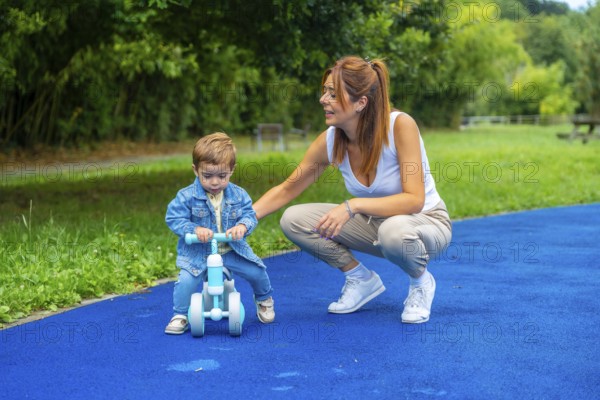 Young mother crouching next to her son helping him learn to ride a balance bike on a blue surface in a park, promoting child development and family bonding