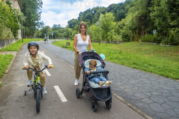 Mother pushing a stroller while walking alongside her son riding a bicycle in a vibrant park, cherishing quality time together outdoors and enjoying the summer sunshine