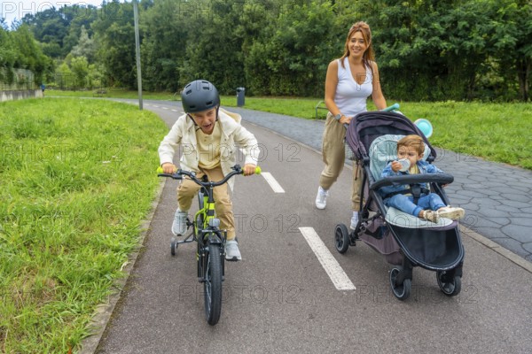 Mother walking with her baby in a stroller while older son riding his bicycle on a paved path, enjoying quality time together outdoors, surrounded by nature and sunlight