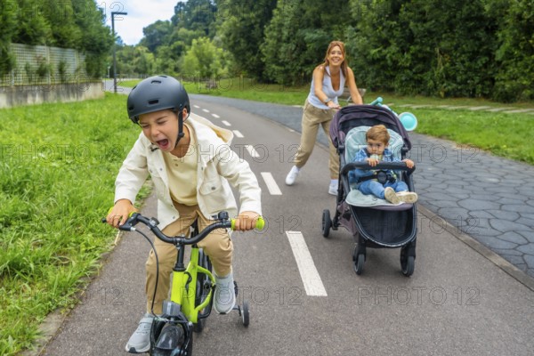Child wearing a helmet is riding a bicycle and shouting, while his mother is pushing a stroller with another child sitting in it, in a park