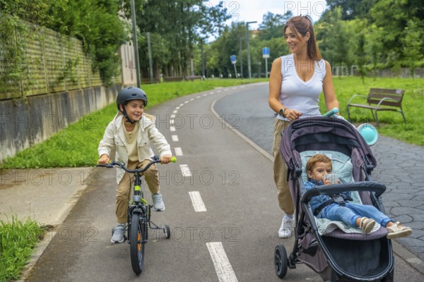 Happy mother walking with her two children, one riding a bicycle with training wheels and the other sitting in a stroller, enjoying a sunny day at the park