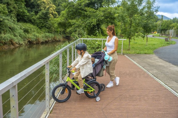 Young mother pushing a stroller with a baby inside and watching her other son riding a bicycle on a wooden walkway next to a river in a park during a sunny summer day