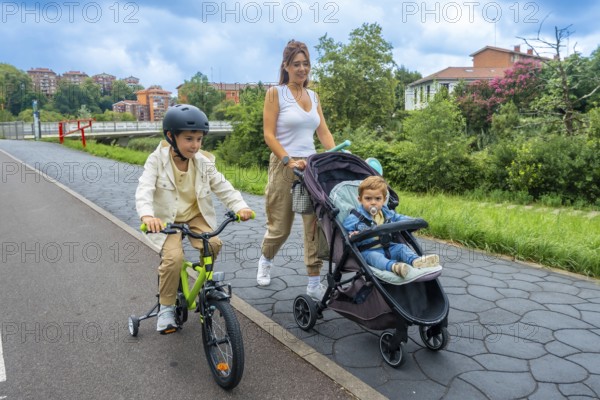 Mother enjoys a leisurely stroll in an urban park, pushing a stroller with her baby while her older son rides alongside on his bicycle, promoting family bonding and an active lifestyle