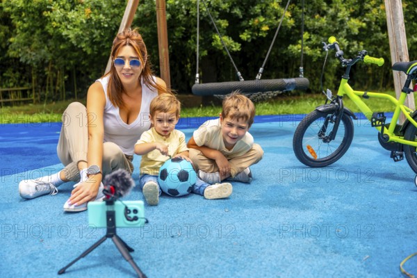 Content creator mother filming video with her sons and a soccer ball on a playground with a swing and a bicycle, using a smartphone, microphone and tripod