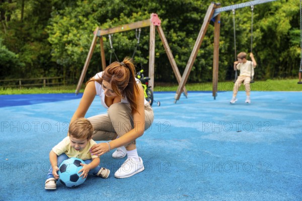 Young mother crouching next to her toddler son playing with a ball on a blue playground surface while another child enjoys swinging on a swing set in the background