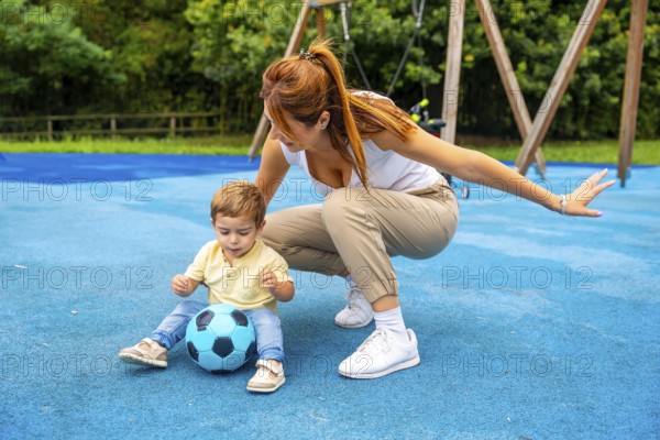 Young mother crouching beside her toddler son, guiding him as he plays with a soccer ball on a vibrant blue playground surface, nurturing early love for sports