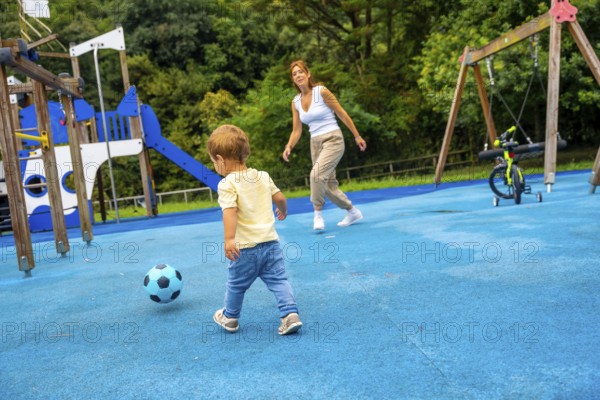 Happy toddler playing soccer with his mother at the playground on a sunny summer day, enjoying quality time together and promoting physical activity