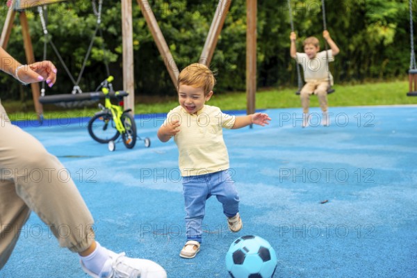 Happy toddler boy kicking soccer ball at playground with family, enjoying a playful moment on a sunny day, creating a joyful and active childhood memory