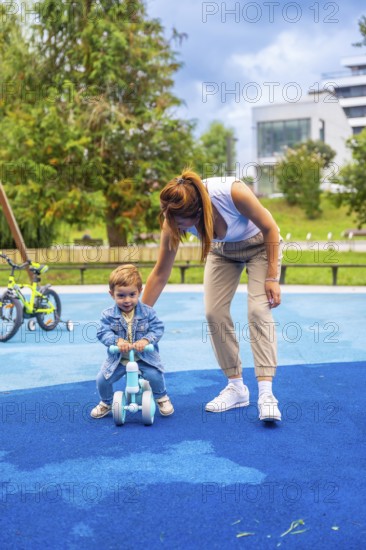 Young mother guiding her son as he learns to ride a balance bike on a playground featuring a soft blue rubberized surface, creating a joyful moment of family bonding and support