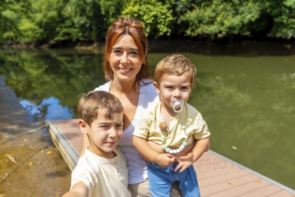 Mother and her two sons are taking a selfie on a wooden dock next to a river, surrounded by lush green trees on a sunny day, creating a heartwarming scene of family bonding in nature