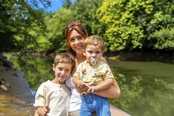 Happy mother holding her baby and posing with her older son by the river in a sunny summer day, enjoying family time together surrounded by nature