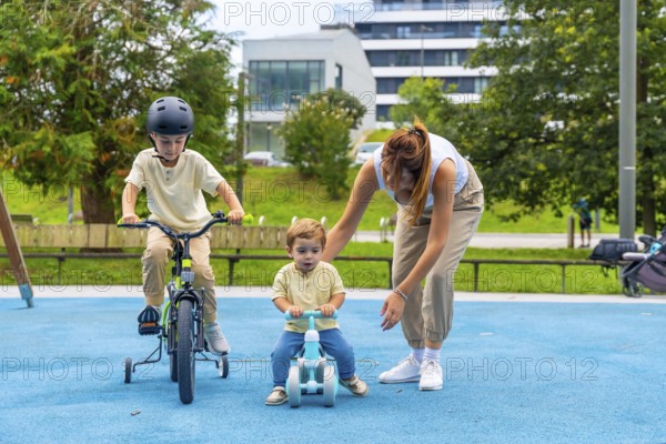 Young mother assisting two children riding bikes in a sunny playground, with the older boy on a bicycle with training wheels and the younger on a balance bike