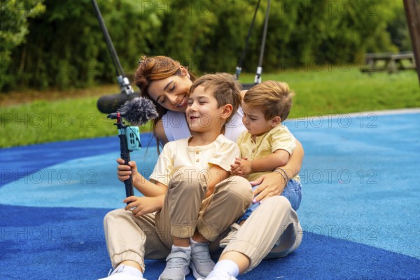Mother and two sons are sitting on playground's rubber surface, holding a smartphone attached to a gimbal with microphone, creating video content for their vlog