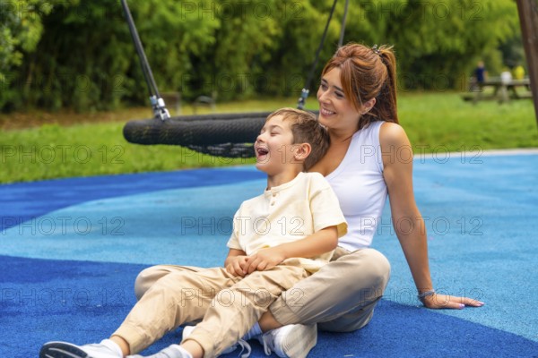 Happy mother and son sitting together on blue rubber flooring near a swing, laughing joyfully and cherishing their playful moments at the vibrant playground
