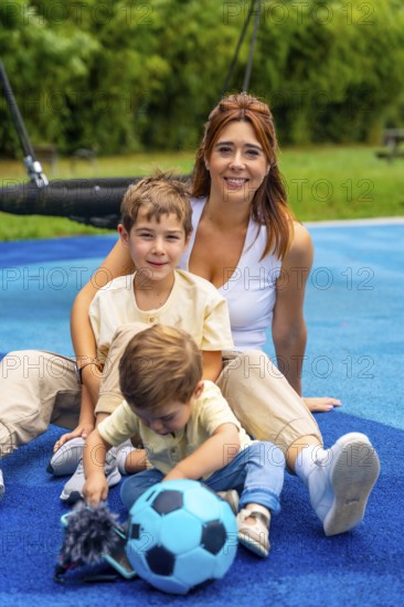 Mother and her two sons are sitting on a playground's blue rubberized surface, enjoying quality time together, with the younger son playing with a light blue soccer ball
