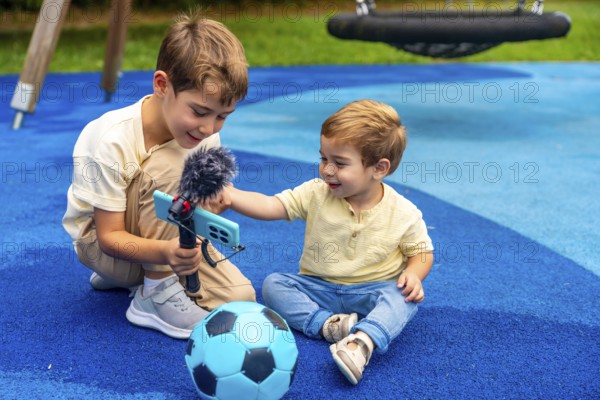Two brothers playing as videobloggers in the playground, capturing fun moments with their phone and microphone while sitting beside a soccer ball, enjoying carefree summer days