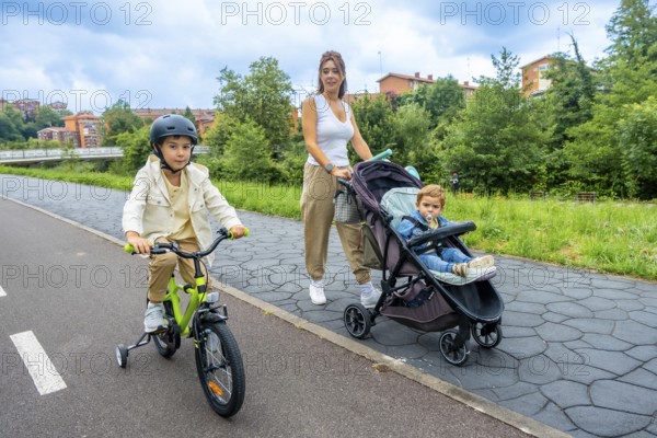 Young mother pushing a stroller with her baby boy, while her older son rides a bicycle with a helmet, enjoying a sunny day filled with laughter and togetherness in the park