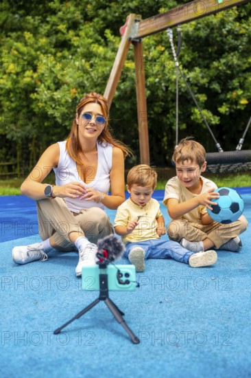 Content creator filming a video with two children at a playground, using a smartphone, microphone, and tripod, while sitting on a blue rubberized surface