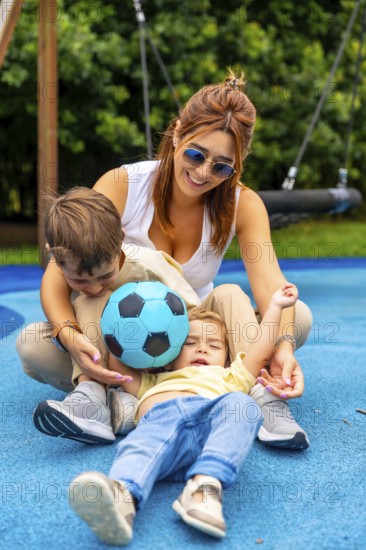 Happy mother and two children playing with a soccer ball on a playground's blue rubberized surface, enjoying quality time together on a sunny day