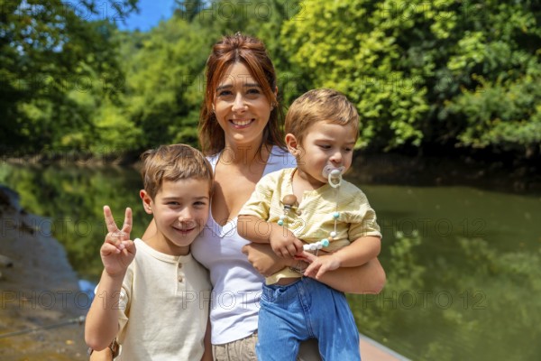 Caucasian mother joyfully holding her two sons near a river on a sunny summer day, with one son making a victory sign, all smiling and engaging with the camera