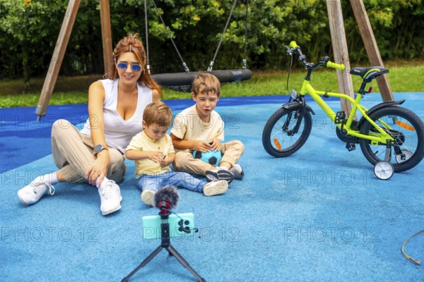 Influencer mother recording a video with her two sons at playground using a smartphone and microphone on a tripod, with a kids bicycle in the background