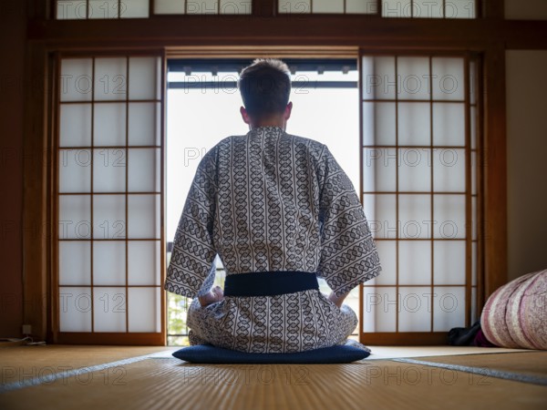 Young man wearing kimono sitting in traditional Japanese living room with tatami mats and shoji sliding doors, from behind, Yamanouchi, Nagano, Japan
