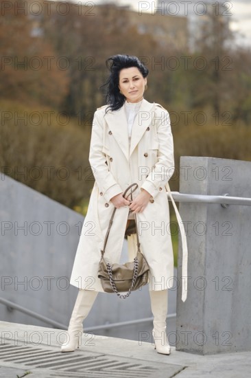 A fashionable woman stands confidently in a city park during autumn. She wears a long cream coat and sleek white boots, showcasing her stylish outfit