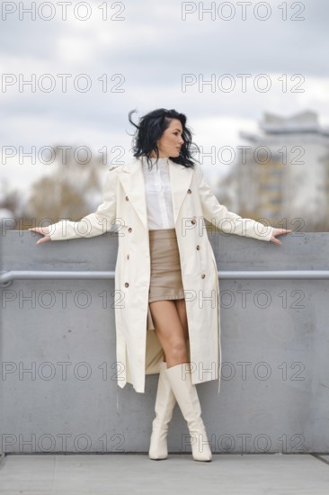 A woman showcases her autumn street style while standing on a balcony. She wears a long white coat over a chic outfit, paired with knee-high boots