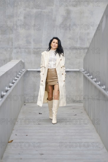 A woman strolls down a sleek walkway, wearing a beige trench coat over a white blouse and a stylish skirt. Her tall boots complement her outfit, set against a modern urban backdrop in autumn