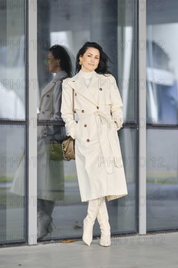 A woman showcases her autumn street style, wearing a cream coat and boots while leaning against a glass building. The scene reflects a stylish urban background, perfect for a fall day