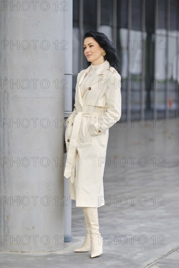 A confident woman stands next to a concrete pillar, dressed in a cream trench coat and matching knee-high boots
