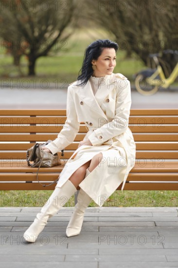 A woman dressed in a stylish cream trench coat and knee-high boots sits gracefully on a wooden bench in a peaceful park