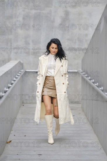 A young woman walks confidently down a sleek concrete staircase, wearing a light-colored trench coat, stylish leather mini skirt, and knee-high boots