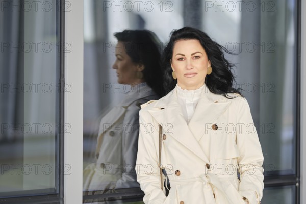 A woman stands confidently in a white trench coat, with her hair elegantly flowing on the wind. She poses near a large glass window reflecting the surrounding urban scenery during autumn
