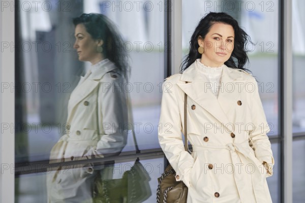 A woman dressed in a chic trench coat stands confidently beside a reflective glass wall. Her long hair flows gently as she poses in a vibrant urban setting, embracing the autumn season