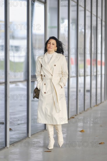 A stylish woman strolls along a sleek urban walkway. She wears a light trench coat and matching knee-high boots, embodying autumn fashion. The backdrop features large glass windows and fallen leaves