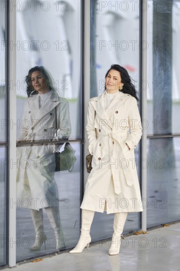 Dressed in a stylish white trench coat and matching boots, a woman stands confidently against a backdrop of glass wall of an urban building
