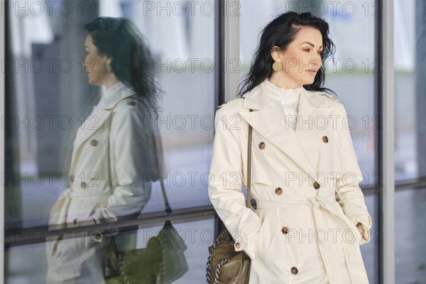 A woman dressed in a fashionable trench coat stands outside, her reflection visible in the glass wall next to her