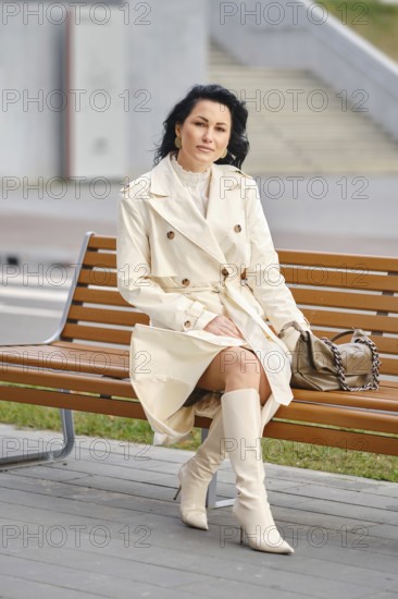 A woman sits on a bench in a city park, wearing a cream trench coat and knee-high boots. Her hair is styled, and she looks relaxed while the golden leaves swirl around her on an autumn day