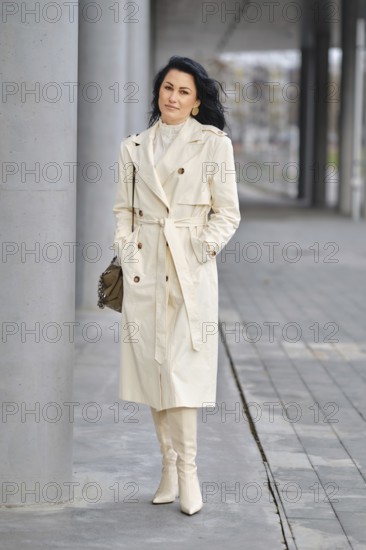 A confident woman strolls along a modern urban walkway, dressed in a light trench coat and matching boots. Her sleek look blends effortlessly with the autumn scenery as she enjoys her day
