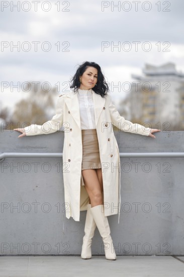A woman stands confidently against a gray barrier, wearing a stylish long coat, knee-high boots, and a skirt. Soft autumn colors surround her as she poses in a tranquil urban environment