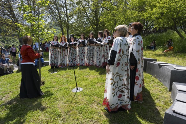 Women's choir in white colorful robes singing outdoors, surrounded by trees and audience, choir at Tatev Monastery, Syunik Province, Syunik, Armenia
