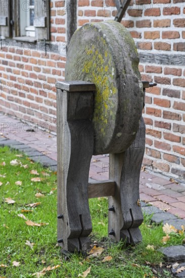 Old whetstone on wooden stand surrounded by grass and brick buildings, Münsterland, North Rhine-Westphalia, Germany