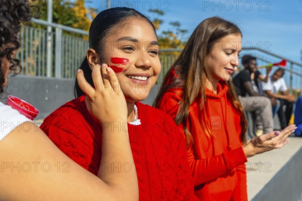 Young happy spectators sitting in bleachers having face paint applied before a sports event, showing team spirit and fan enthusiasm in a sunny outdoor stadium
